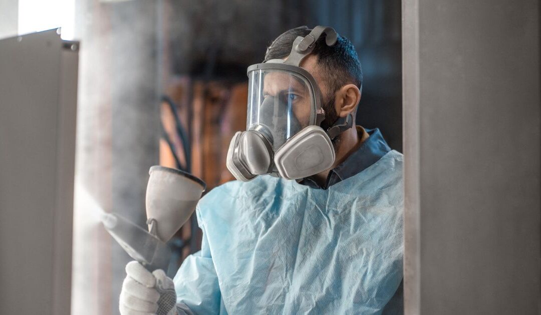 a person in protective gear (mask, gloves, etc.) spraying powder coating materials